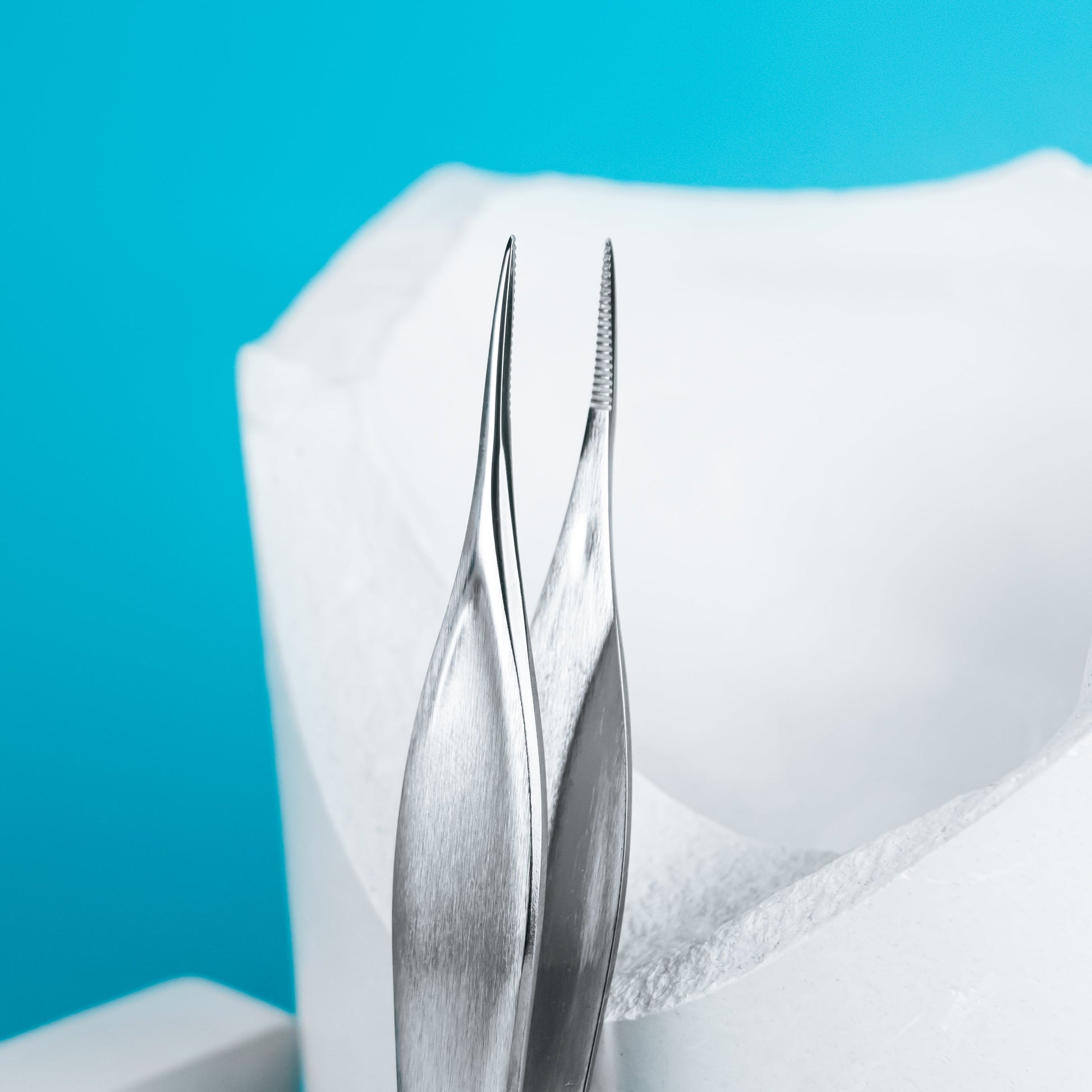 A close-up of two rust-resistant STALEKS PRO Podology Splinter Tweezers PODO 10 by Staleks, with pointed tips, displayed on a bright blue background next to a white dental model—ideal for ingrown nail care or podology tasks.