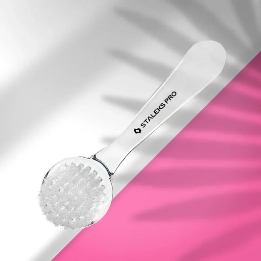 The STALEKS PRO Manicure Brush EXPERT 01 For Dust Removal (With Cover) by Staleks, featuring a clear handle and white bristles, rests on a diagonally split pink and white background with soft leafy shadows.