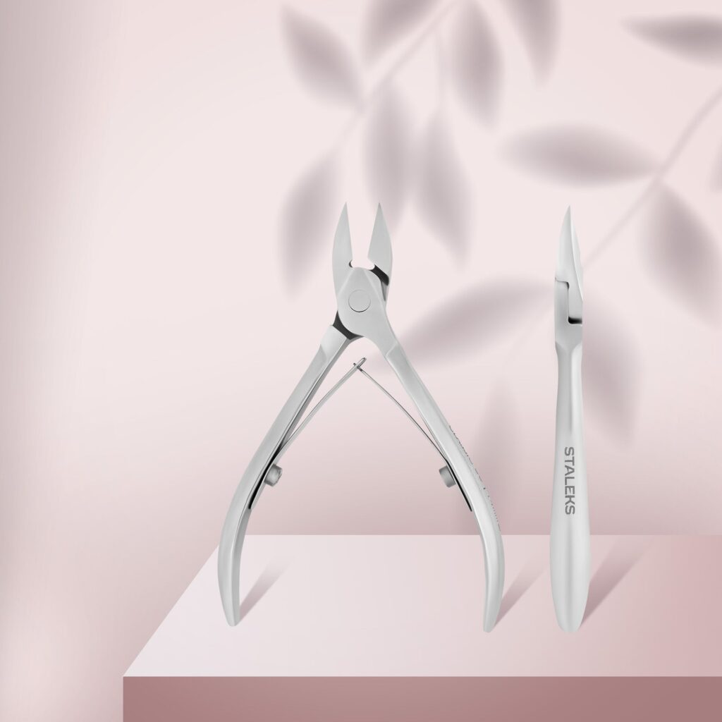 Two stainless steel manicure tools—a STALEKS PRO Nail Nippers CLASSIC 63 14 mm and a cuticle pusher—stand upright on a pink surface with soft leaf shadows. The nipper on the right is branded "Staleks.