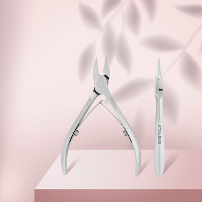 Two stainless steel manicure tools—a STALEKS PRO Nail Nippers CLASSIC 63 14 mm and a cuticle pusher—stand upright on a pink surface with soft leaf shadows. The nipper on the right is branded "Staleks.