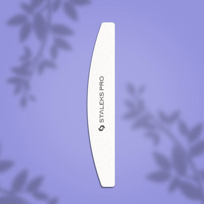 A white STALEKS PRO Grinding Buff crescent EXPERT 41 100/180 grit nail file (Staleks) lies vertically on a purple background with blurred leaf silhouettes, highlighting its fine abrasive surface.