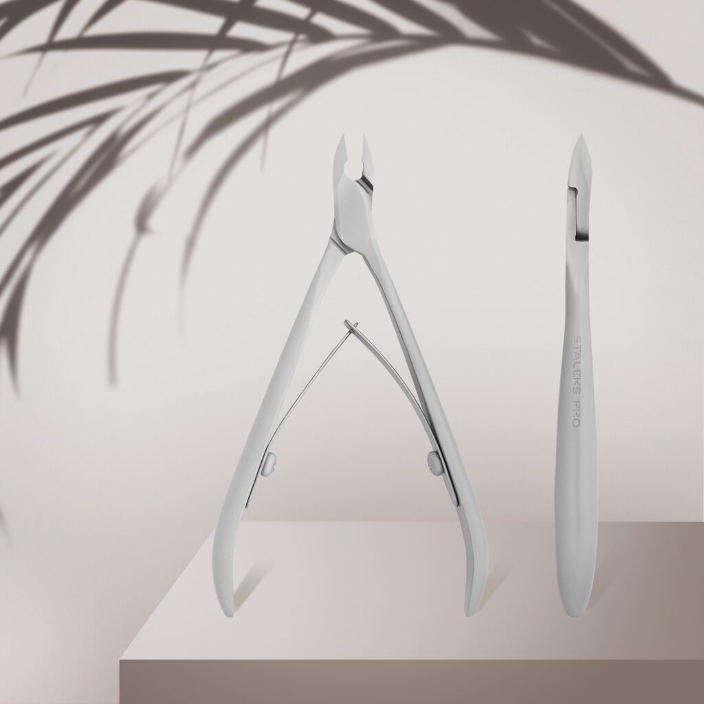 The Staleks STALEKS PRO Professional Cuticle Nippers SMART 11 7 mm, with a satin finish, and a cuticle pusher are displayed on a beige podium, accented by the soft shadow of leafy foliage in the background.