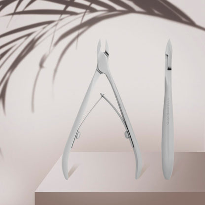 The Staleks STALEKS PRO Professional Cuticle Nippers SMART 11 7 mm, with a satin finish, and a cuticle pusher are displayed on a beige podium, accented by the soft shadow of leafy foliage in the background.