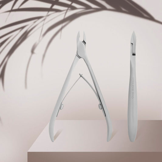 The Staleks STALEKS PRO Professional Cuticle Nippers SMART 11 7 mm, with a satin finish, and a cuticle pusher are displayed on a beige podium, accented by the soft shadow of leafy foliage in the background.