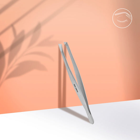 The Staleks PRO Eyebrow Tweezers CLASSIC 10 TYPE 3 (wide beveled) stand upright on a white surface against a peach background, with leaf shadows and an eyelash logo—ideal for shaping thick brows.