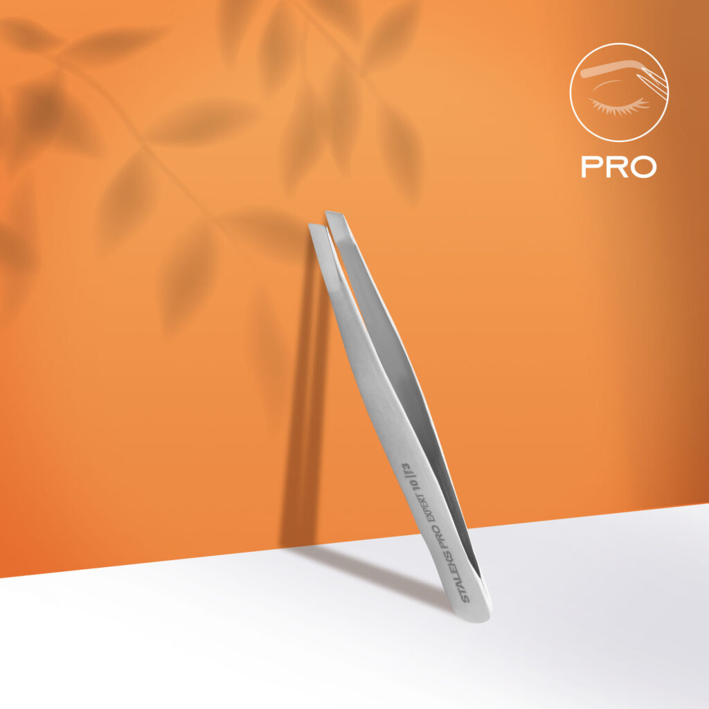 A pair of Staleks PRO Eyebrow Tweezers EXPERT 10 TYPE 3 (wide beveled), made of stainless steel, stands upright on a white surface with leaf shadows and an orange backdrop. The stylized eye logo and "PRO" appear in the top right corner.