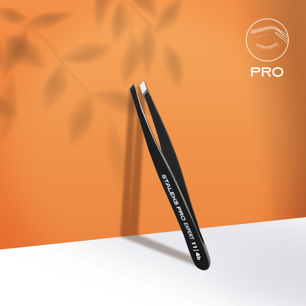 The STALEKS PRO Eyebrow Tweezers Black EXPERT 11 TYPE 4b (Narrow Beveled) by Staleks stand upright on an orange background with leaf shadows, ideal for eyebrow correction; a white eyelash logo appears in the top right corner.