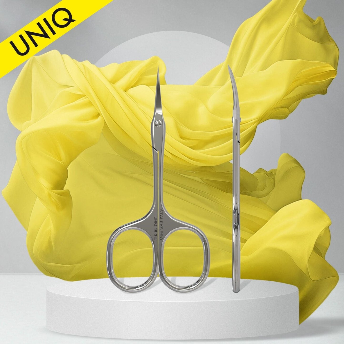 The Staleks STALEKS PRO Professional Cuticle Scissors Ballerina UNIQ 10 TYPE 3 are shown upright and sideways on a white platform, with a flowing yellow fabric backdrop and a "UNIQ" yellow banner in the top left corner.
